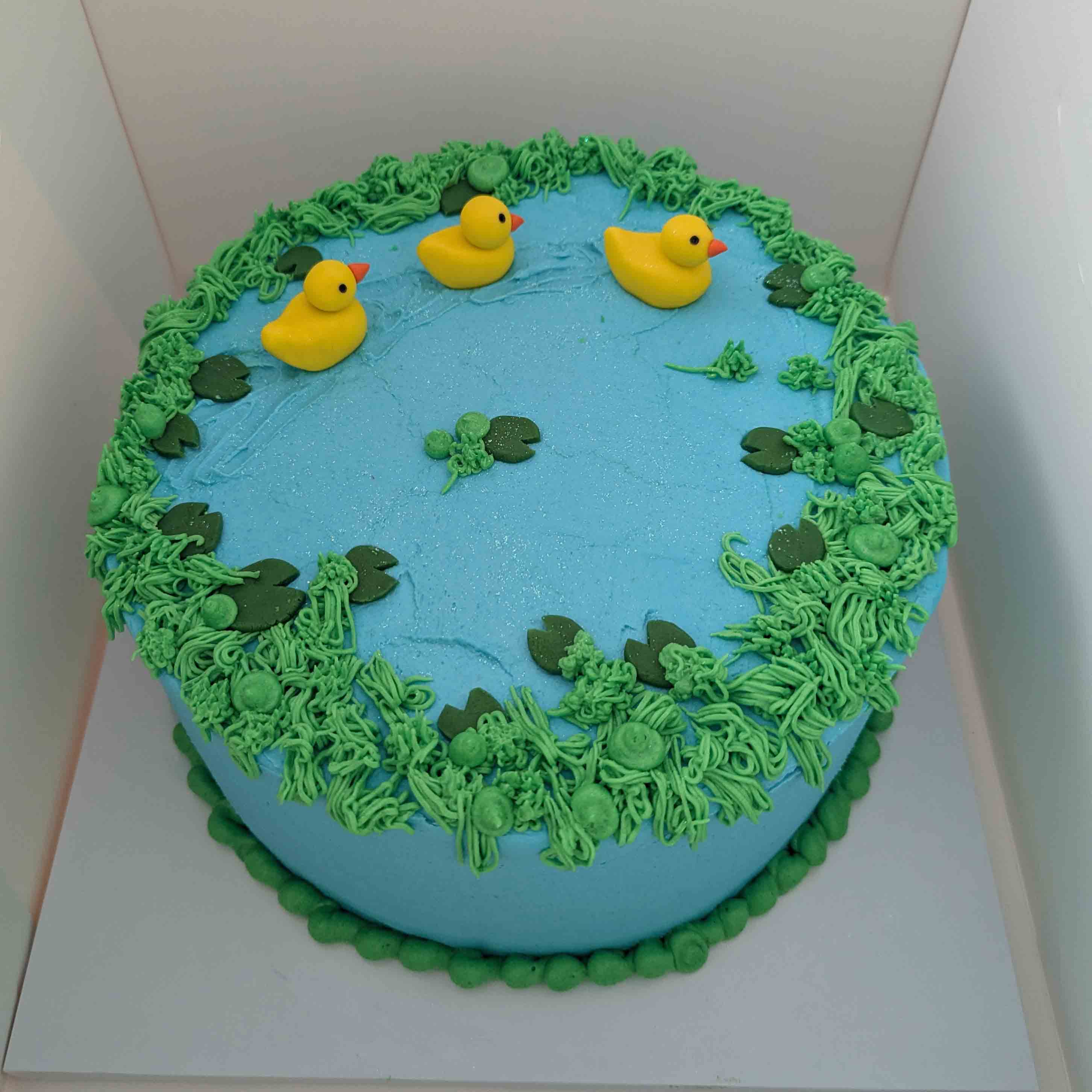 Blue pond-themed cake with three yellow rubber ducks and piped buttercream lily pads